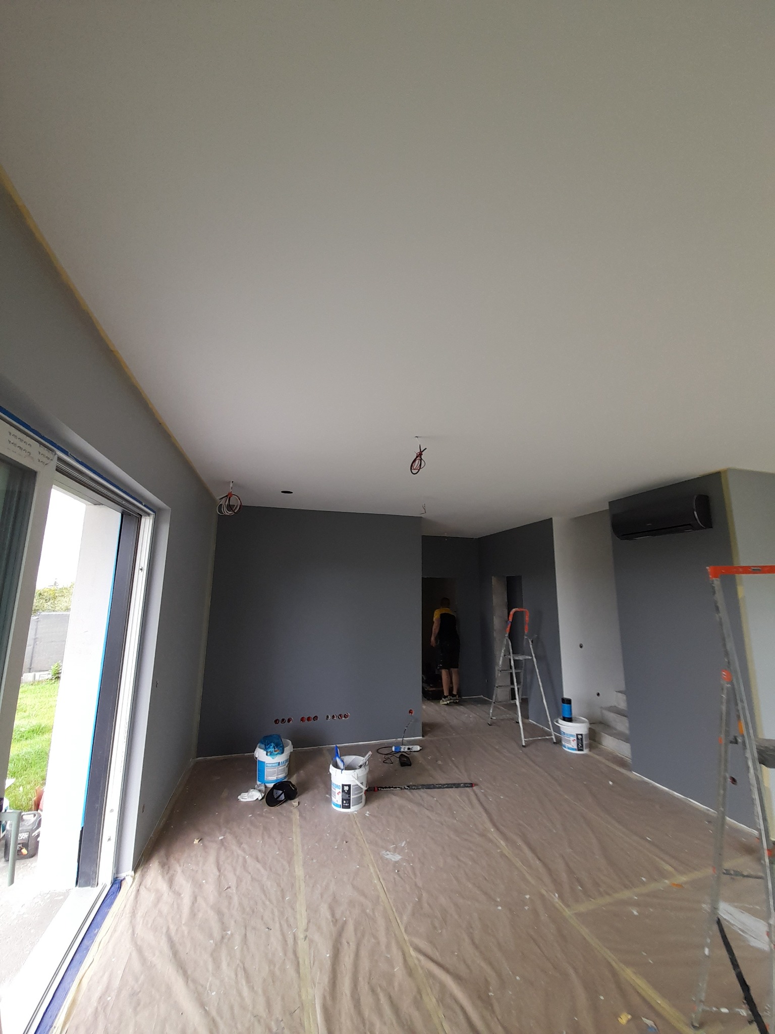 Wnętrze w Radziszewie w trakcie malowania na szaro, widoczne drabiny, wiaderka z farbą, zabezpieczona podłoga folią malarską oraz osoba w oddali.