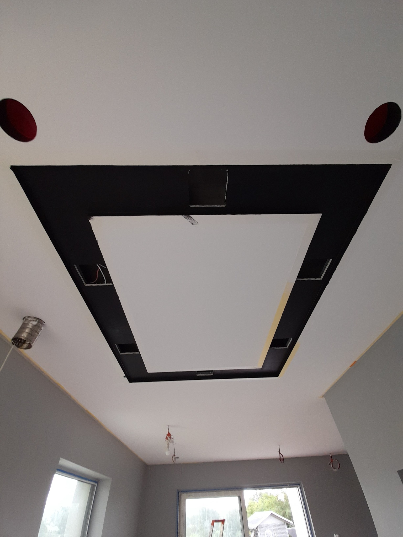 Wielopoziomowy sufit podwieszany z czarnymi elementami, otwory na oświetlenie i widoczne przewody elektryczne, w trakcie montażu w szarym pomieszczeniu z oknami.