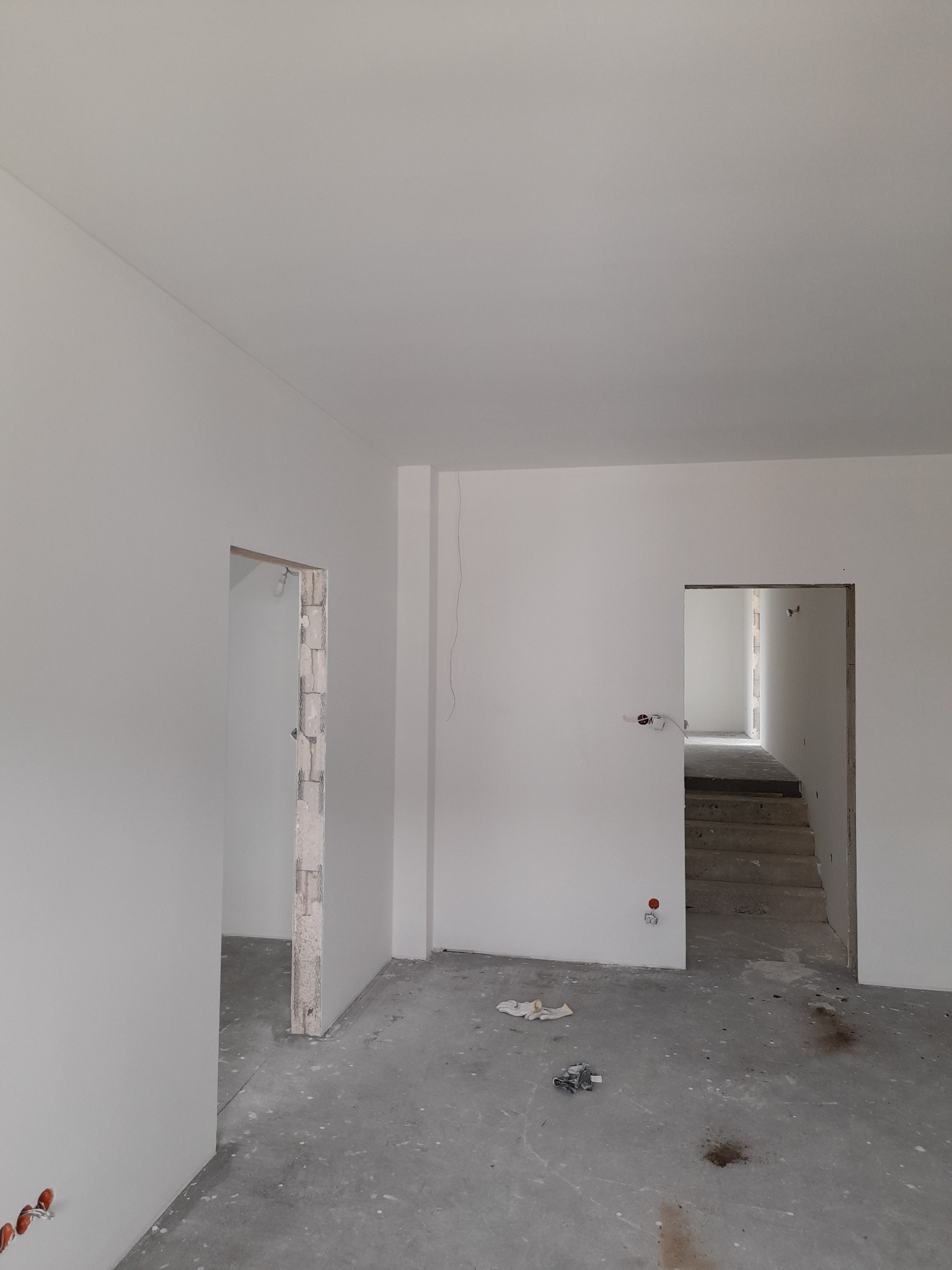 Surowe wnętrze w trakcie wykańczania: białe ściany, surowy beton na podłodze, widoczne instalacje elektryczne i otwory drzwiowe z odsłoniętą cegłą, schody w głębi.