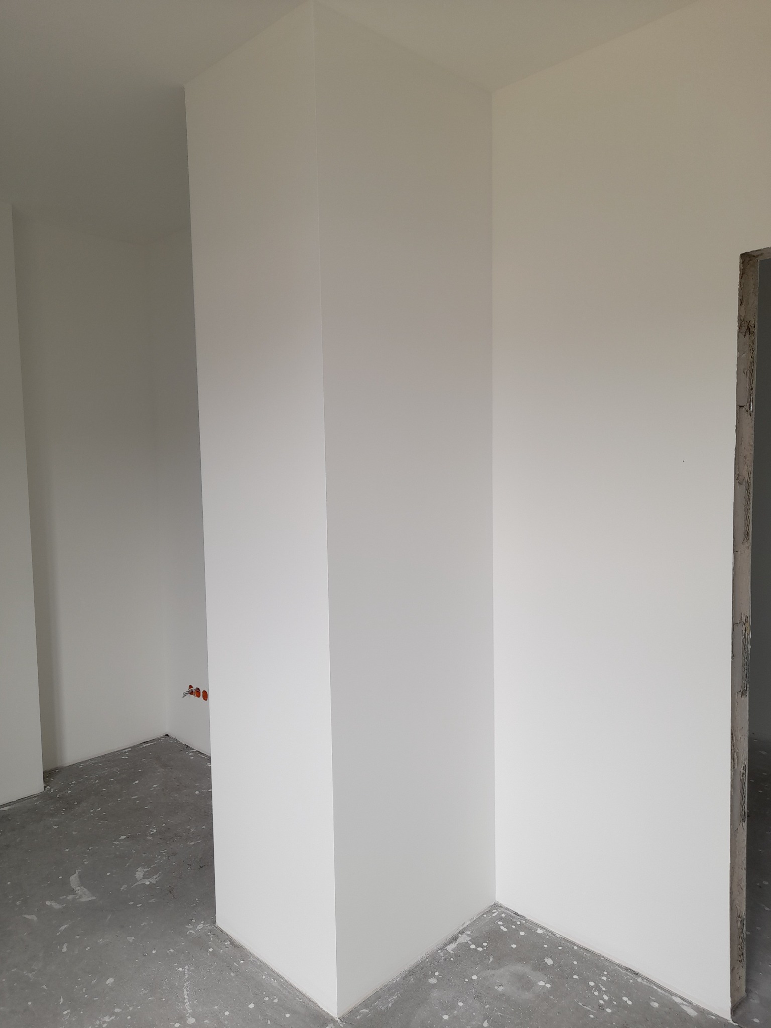 Wnętrze w trakcie wykańczania: świeżo pomalowane, białe ściany, surowy beton na podłodze z widocznymi plamami farby, oraz fragment surowego otworu drzwiowego.