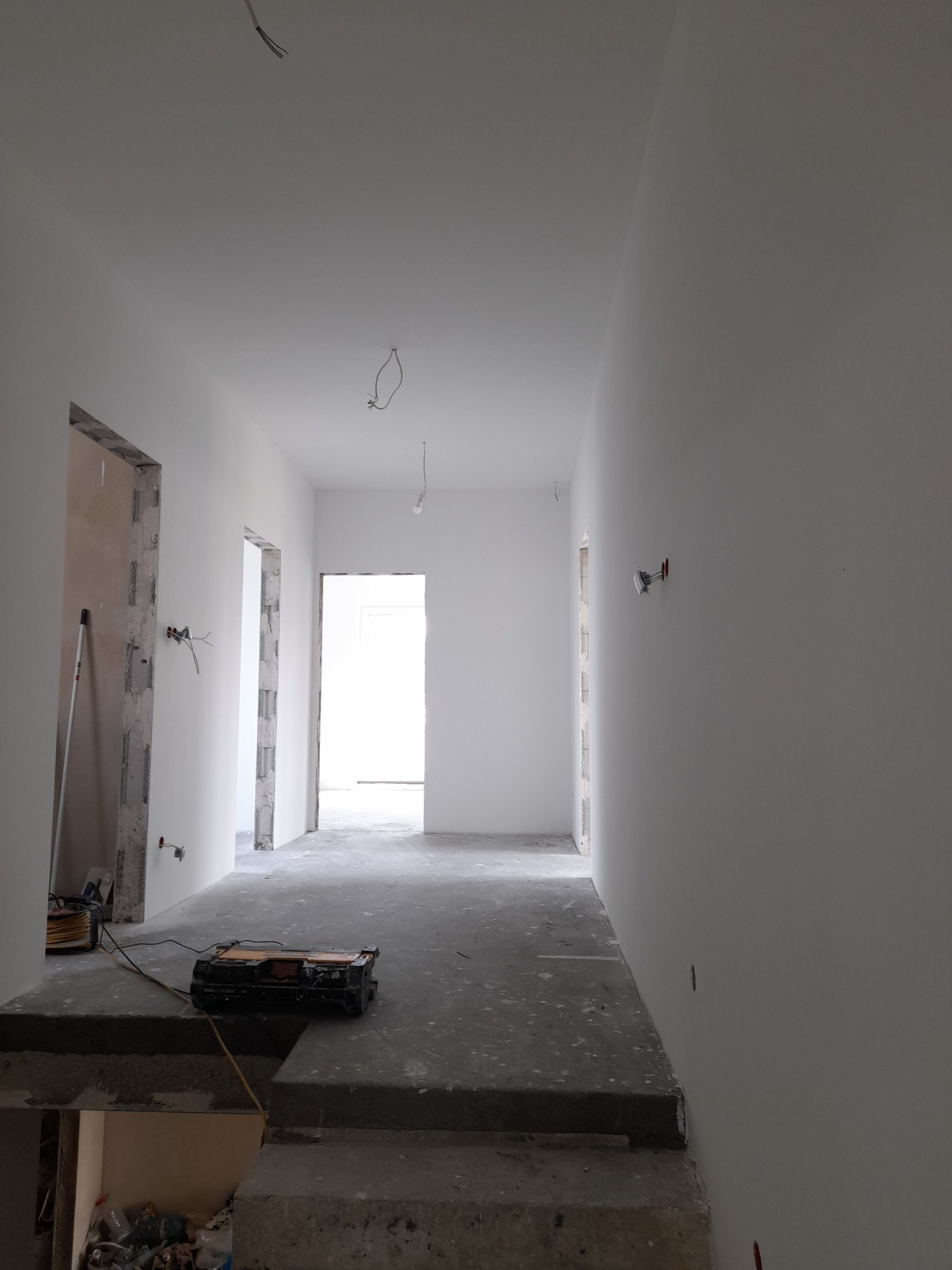 Wnętrze w trakcie prac wykończeniowych: świeżo pomalowane na biało ściany, surowy beton na podłodze, widoczne przewody elektryczne i narzędzia budowlane na podwyższeniu.