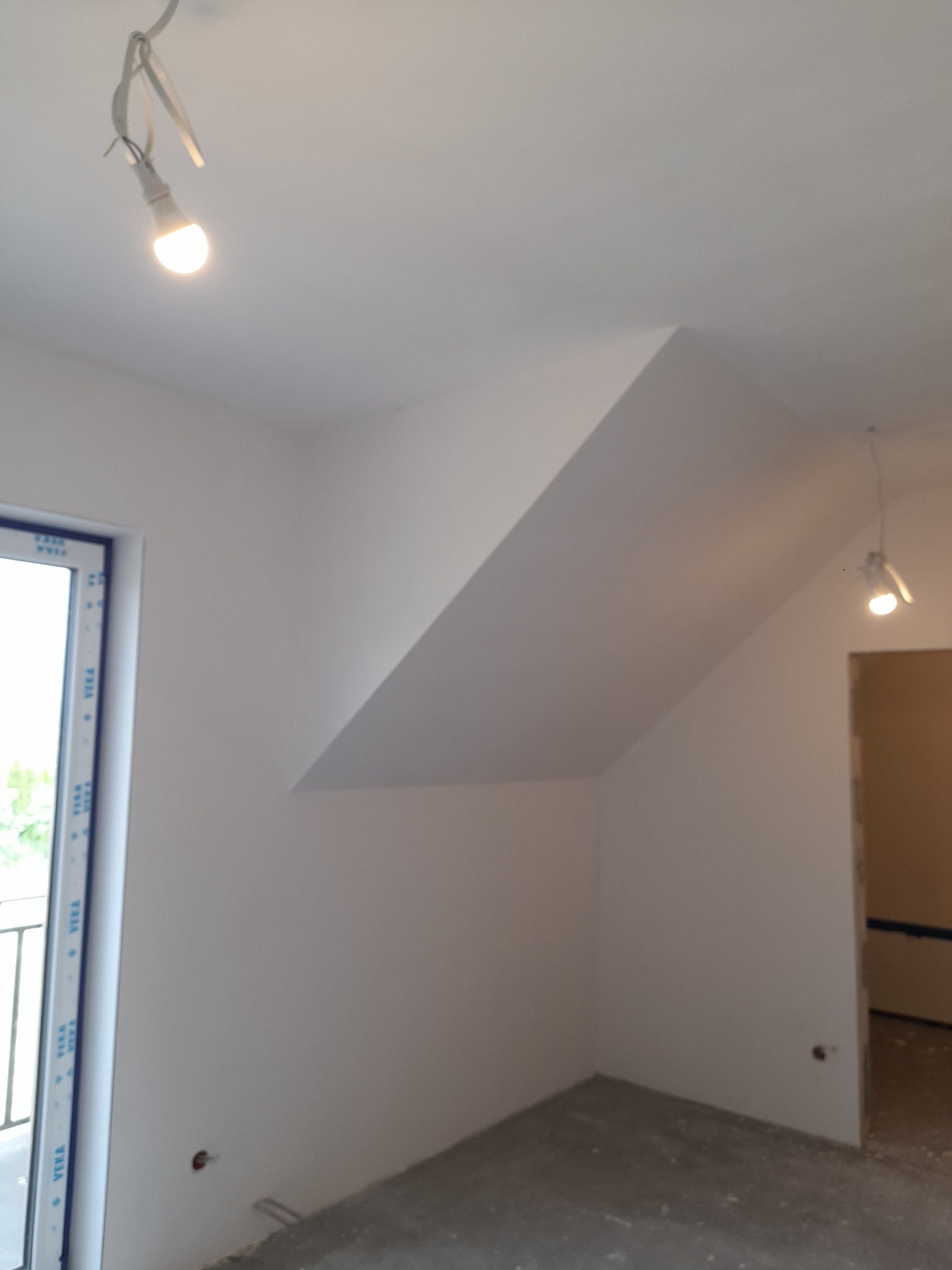 Pomalowane na biało poddasze ze skosami, widoczne okno balkonowe z niebieskimi ramami, betonowa podłoga i wiszące lampy z żarówkami LED.