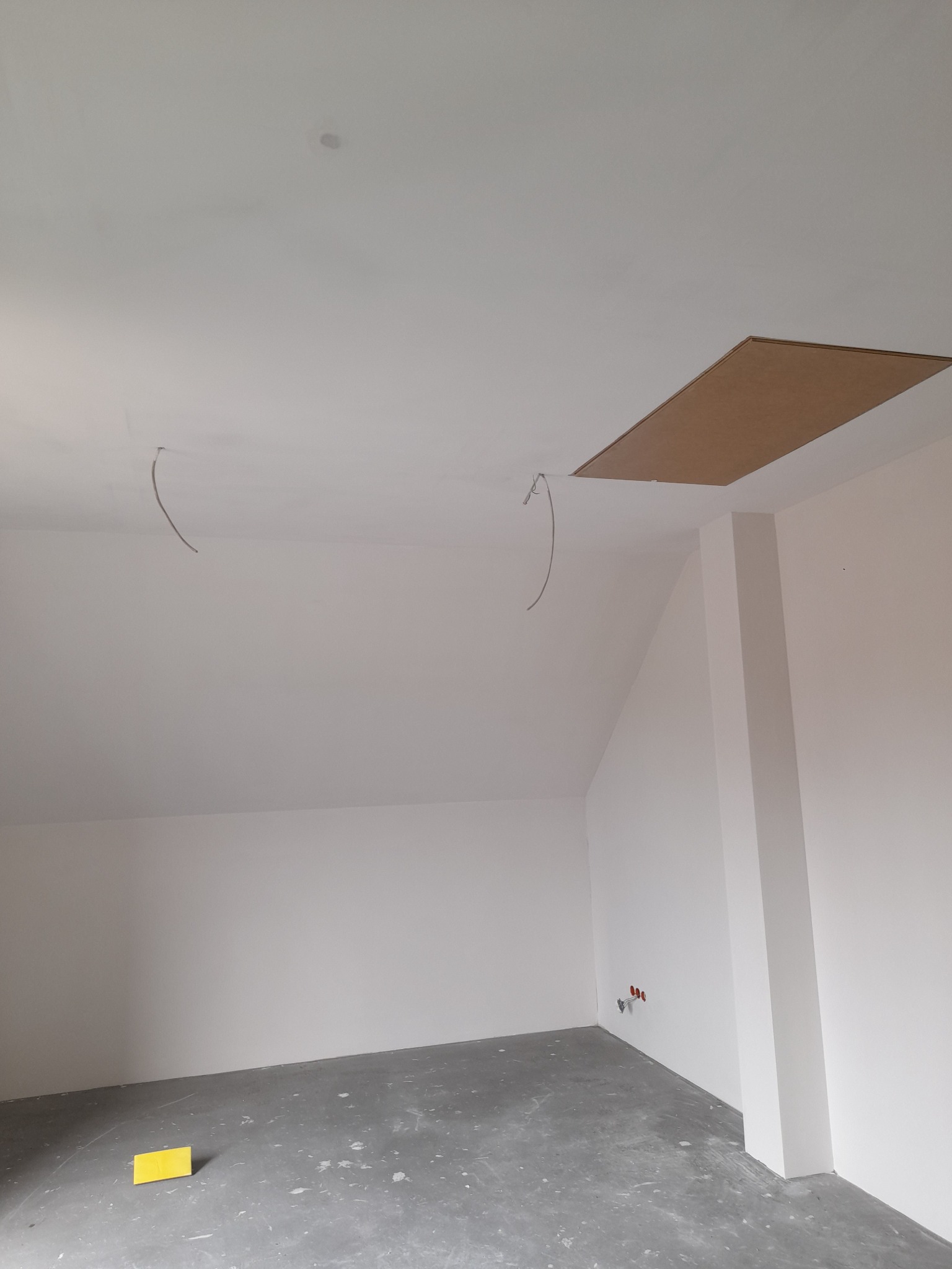 Wnętrze pokoju na poddaszu w trakcie wykańczania, widoczne białe ściany, surowa podłoga, otwór w suficie z drewnianą klapą i wystające przewody elektryczne.