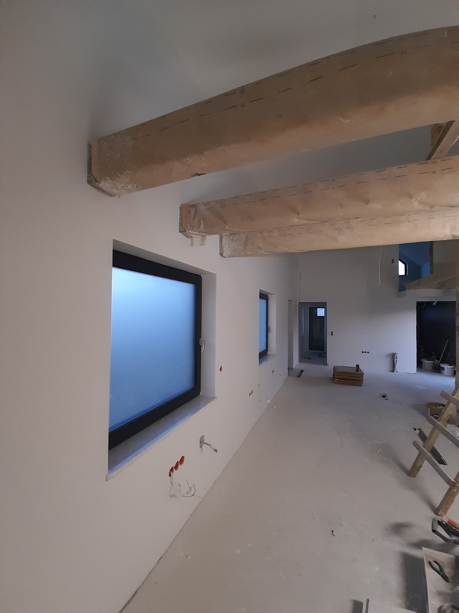 Wnętrze w trakcie wykańczania: białe ściany, okna z czarnymi ramami, zabezpieczone drewniane belki stropowe, widoczne instalacje elektryczne i drabina w tle.