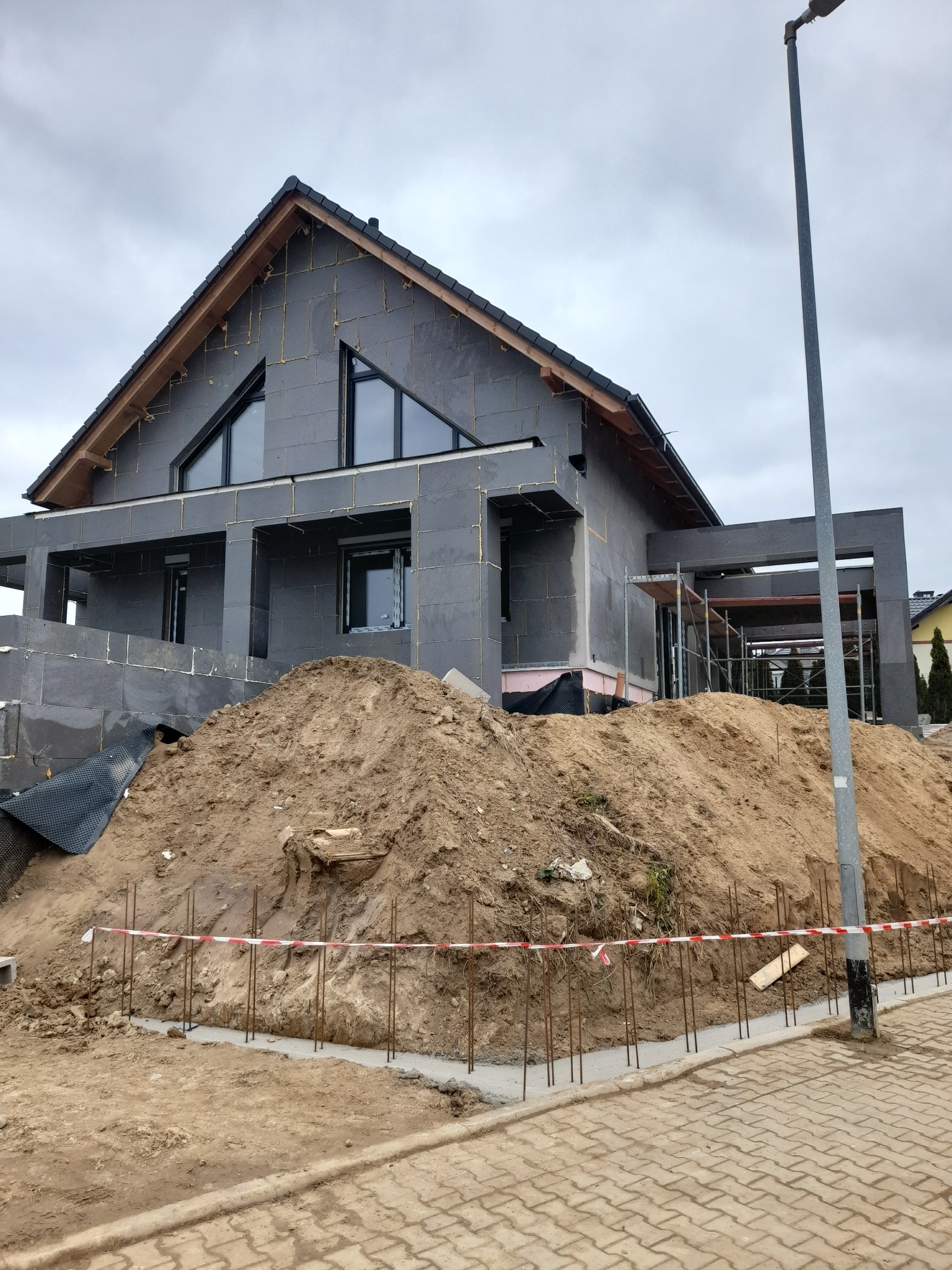 Dom jednorodzinny w trakcie ocieplania ścian zewnętrznych, widoczna warstwa izolacyjna z szarego styropianu, okna z trójkątnymi nadświetlami, ziemia usypana wokół fundamentów.