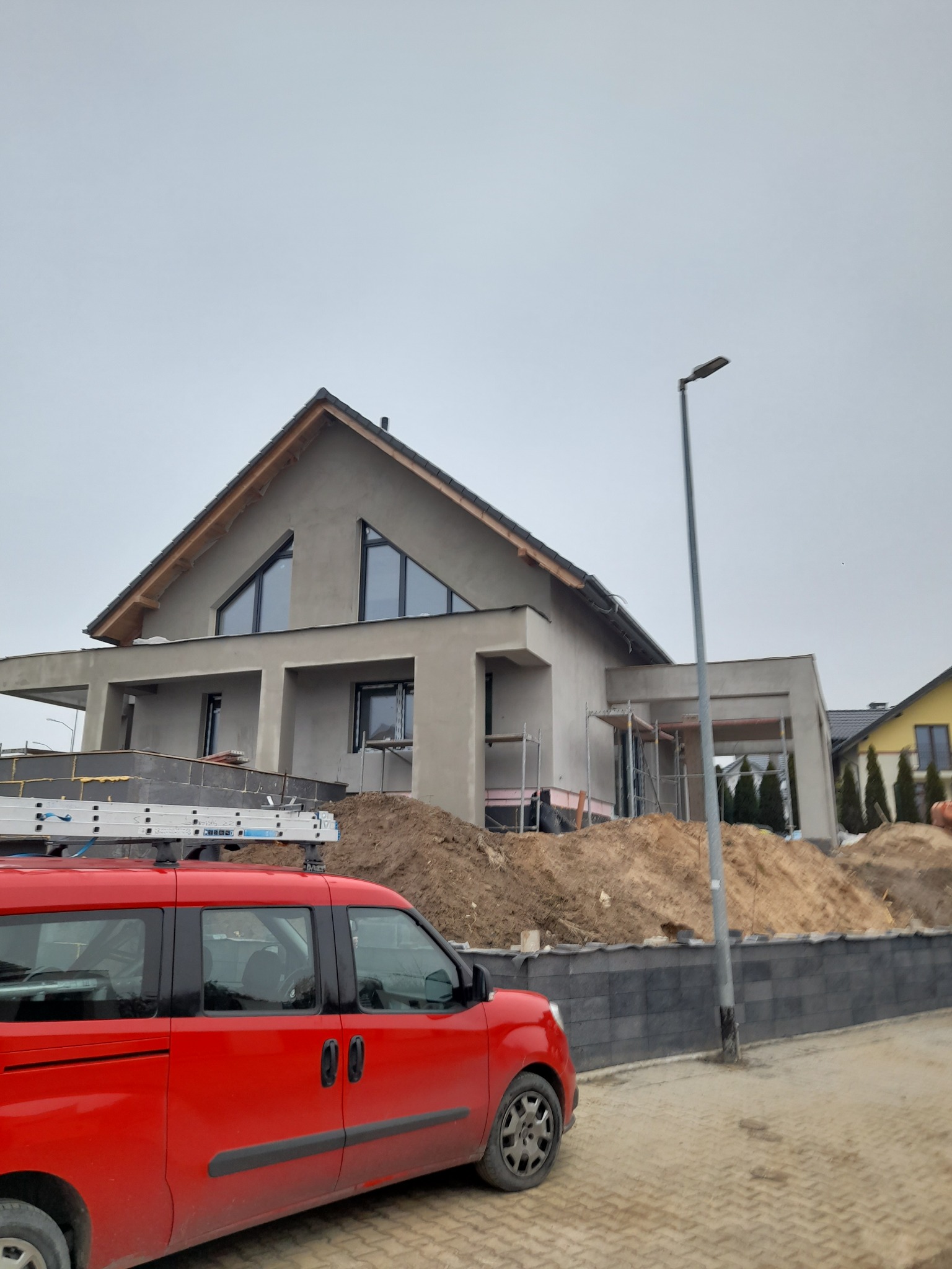 Nowoczesny dom jednorodzinny w trakcie wykańczania elewacji, widoczny czerwony samochód z drabiną na dachu i sterty ziemi przed budynkiem.