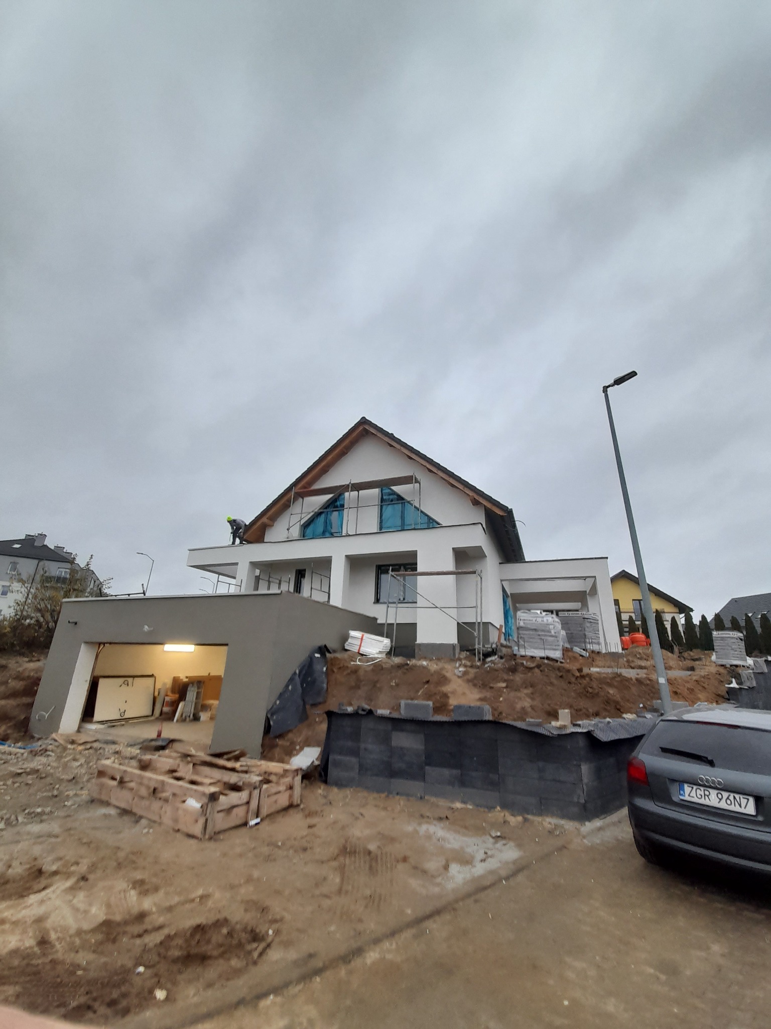 Budowa nowoczesnego domu jednorodzinnego z garażem w trakcie realizacji, widoczny pracownik na dachu, materiały budowlane i samochód osobowy w pobliżu.