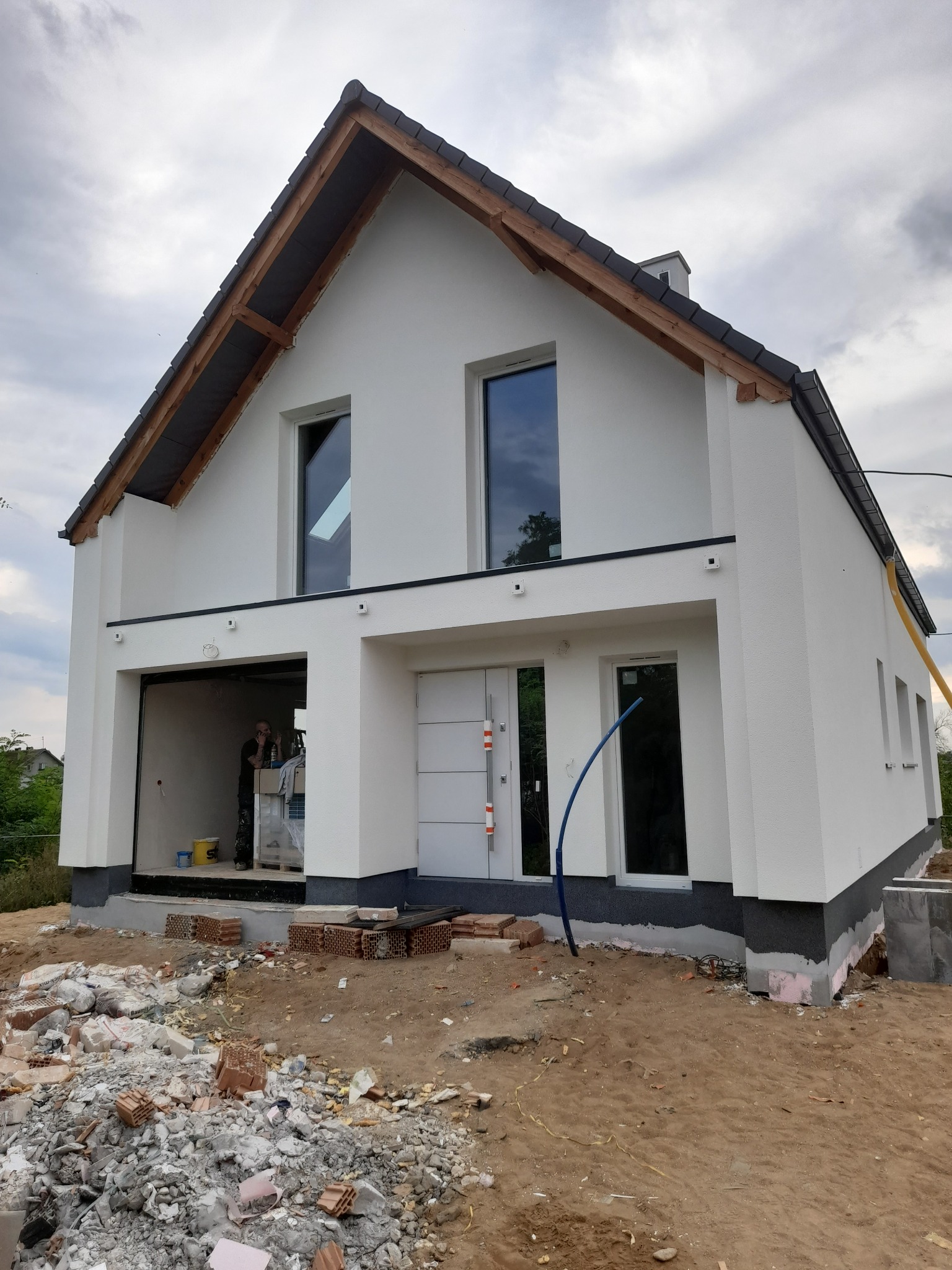 Nowoczesny dom jednorodzinny z białymi ścianami i grafitowym dachem w trakcie wykończenia, widoczne materiały budowlane i pracownik w garażu.