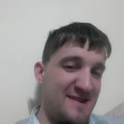 Uśmiechnięty mężczyzna w koszuli w paski, z krótką fryzurą, pozujący na tle ściany z obrazem.