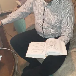 Mężczyzna siedzi po turecku z otwartą książką na kolanach, w tle widoczny laptop na stoliku kawowym i fragment sofy.