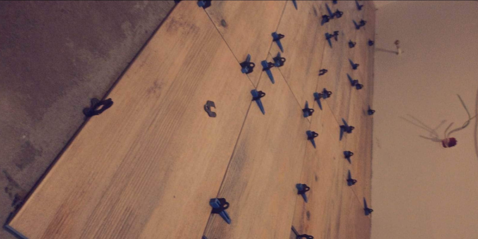 Proces układania płytek imitujących drewno na ścianie z użyciem niebieskich klipsów poziomujących i sznurka, widoczne przewody elektryczne w tle.