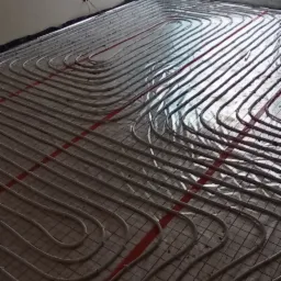 Białe rury grzewcze ułożone w pętle na podłodze z siatką i folią izolacyjną, widok na okno z niebieską folią ochronną i krajobraz za oknem.