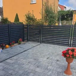 Nowoczesne, poziome, szare przęsła ogrodzeniowe i brama wjazdowa na posesji z kostki brukowej, z dekoracyjnymi kwiatami w donicach i dynią, widoczne narzędzia montażowe.