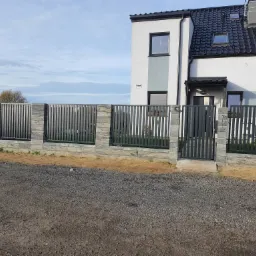 Nowoczesne ogrodzenie z paneli pionowych w kolorze grafitowym, osadzone na niskim murku z szarej kostki, z furtką prowadzącą do domu w tle, widok z zewnątrz.