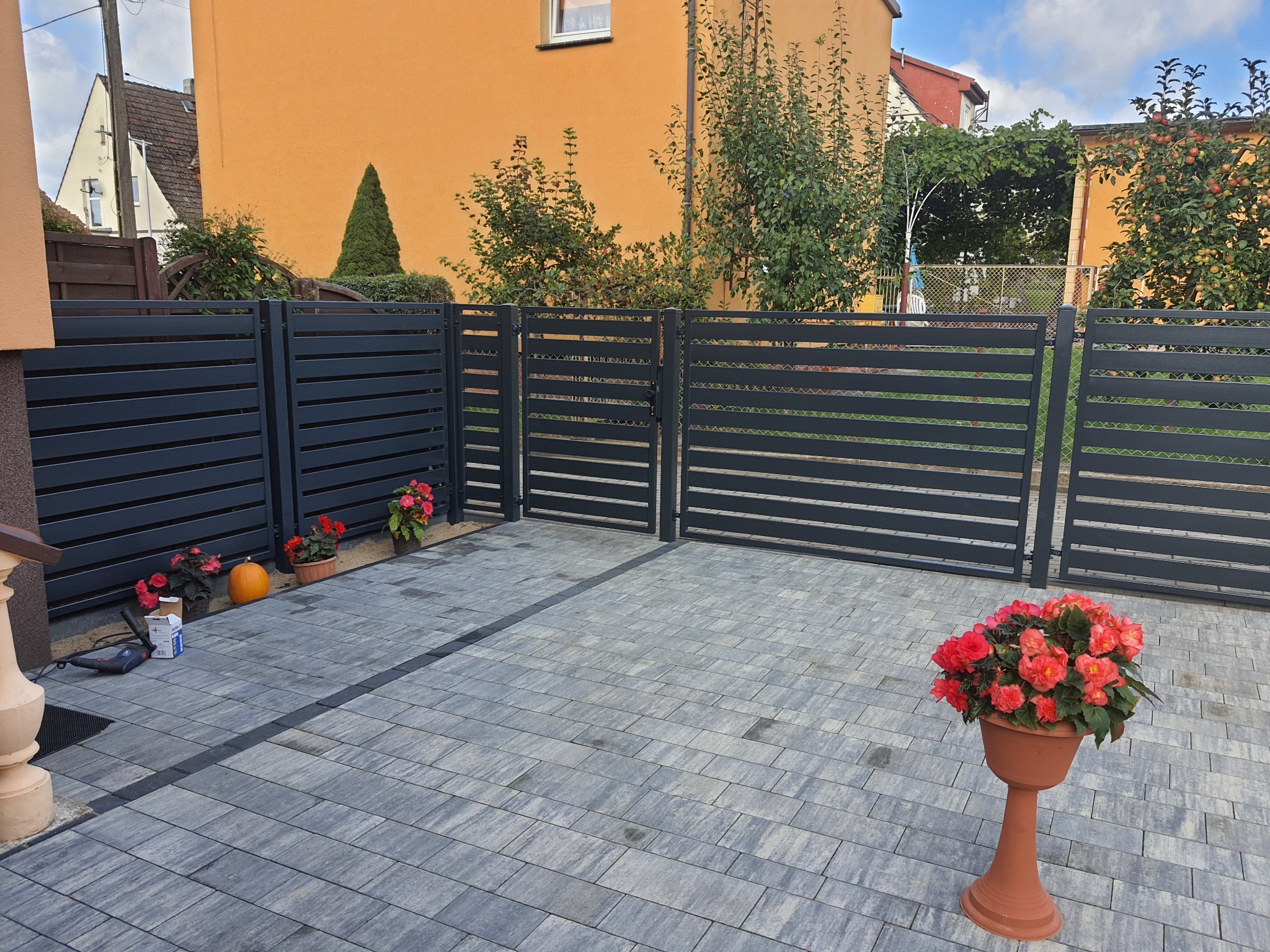 Nowoczesne, poziome, szare przęsła ogrodzeniowe i brama wjazdowa na posesji z kostki brukowej, z dekoracyjnymi kwiatami w donicach i dynią, widoczne narzędzia montażowe.