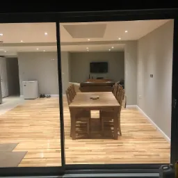 Widok z zewnątrz przez przesuwne szklane drzwi na jasne, nowo pomalowane wnętrze z drewnianą podłogą, stołem i krzesłami; świeżo pomalowane ściany w odcieniach szarości.