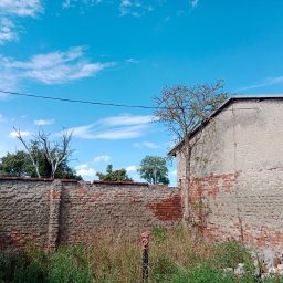 BW-Computers Bogusz Wawrzyniak - Fragment zniszczonego muru z czerwonej cegły i szarego betonu, z widoczną rurą wystającą z ziemi, na tle błękitnego nieba z chmurami.