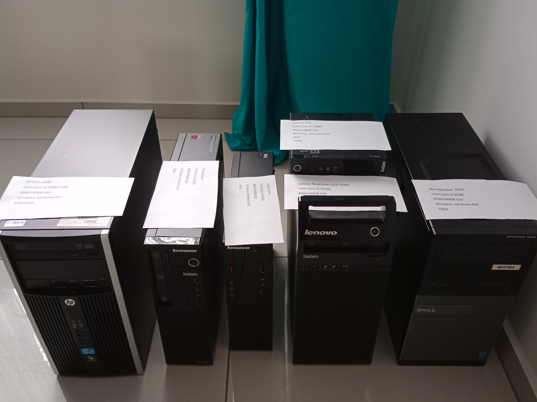 Rząd używanych komputerów stacjonarnych różnych marek (HP, Lenovo, Dell) z naklejonymi specyfikacjami technicznymi, ustawionych na podłodze z zielonym tłem.