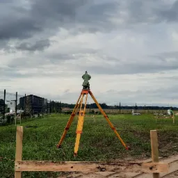 Zielony niwelator optyczny na ż&oacute;łtym statywie na tle pochmurnego nieba i zielonej łąki, z drewnianą konstrukcją w pierwszym planie.
