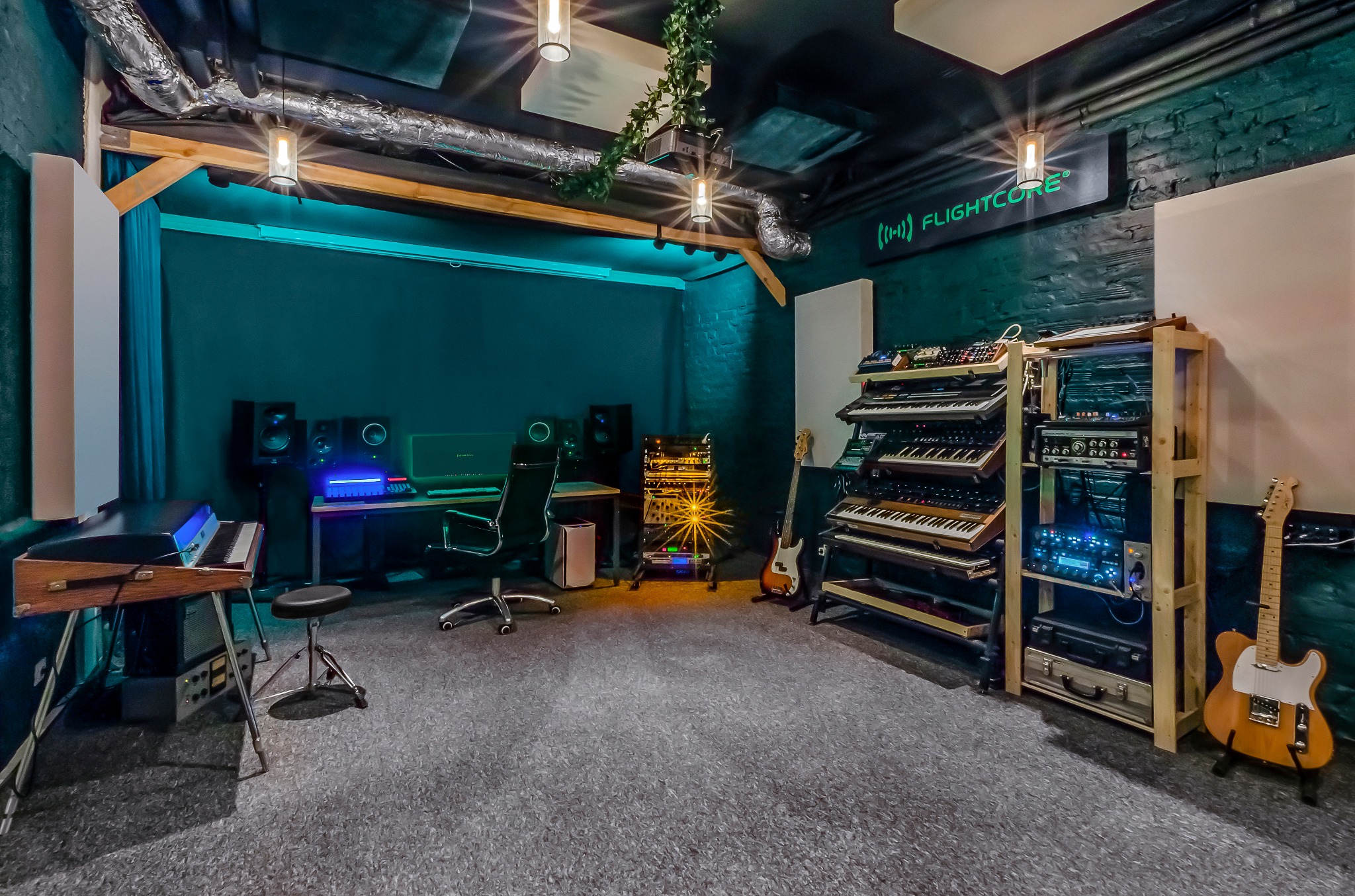 Profesjonalne pomieszczenie z instrumentami muzycznymi, syntezatorami, gitarami i sprzętem audio, oświetlone wiszącymi lampami i neonem z logo 'Flightcore'.