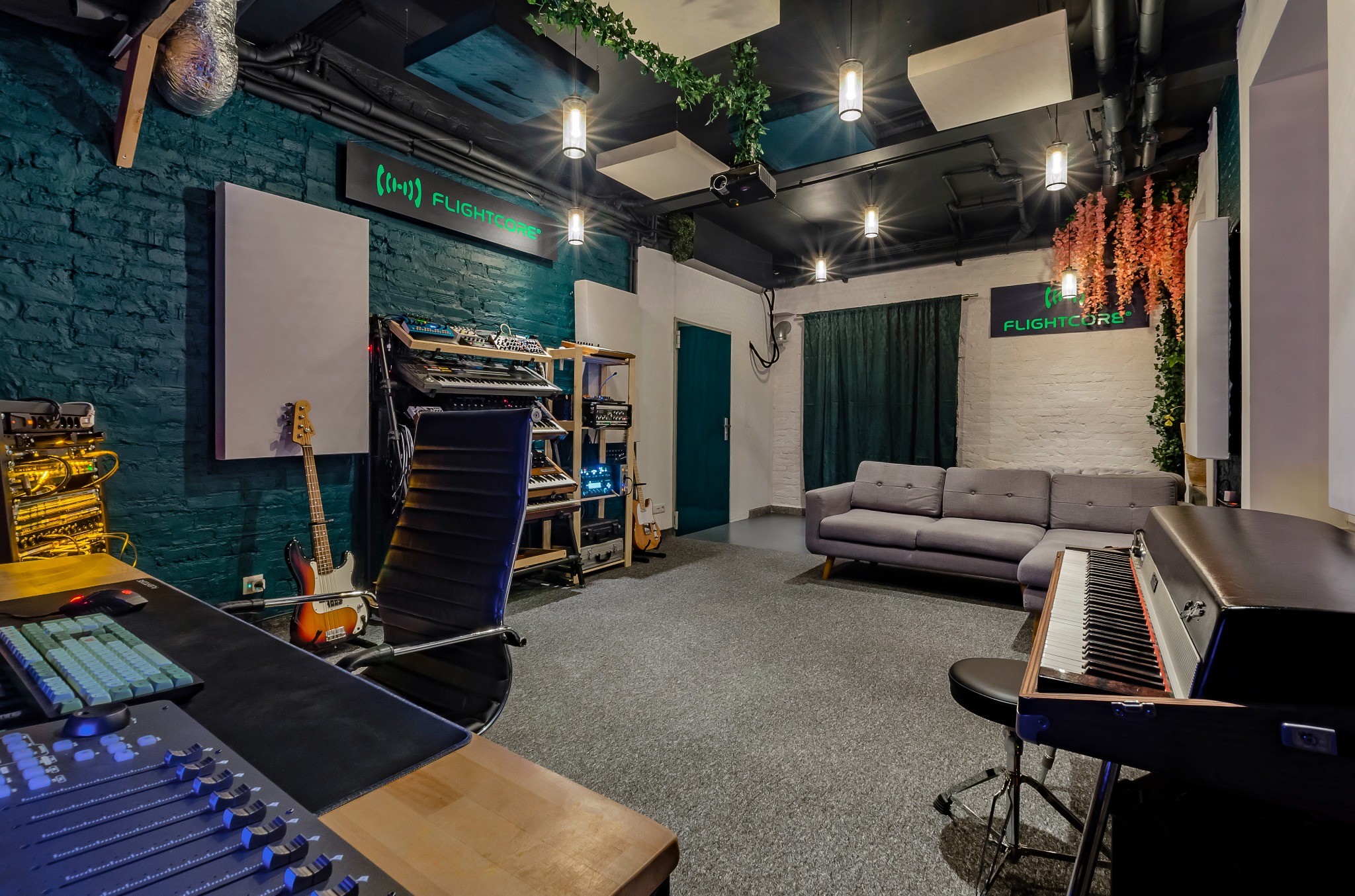 Wnętrze studia nagrań z mikserem, instrumentami klawiszowymi, basem i sofą, oświetlone lampami w stylu industrialnym i ozdobione zielonymi pnączami.