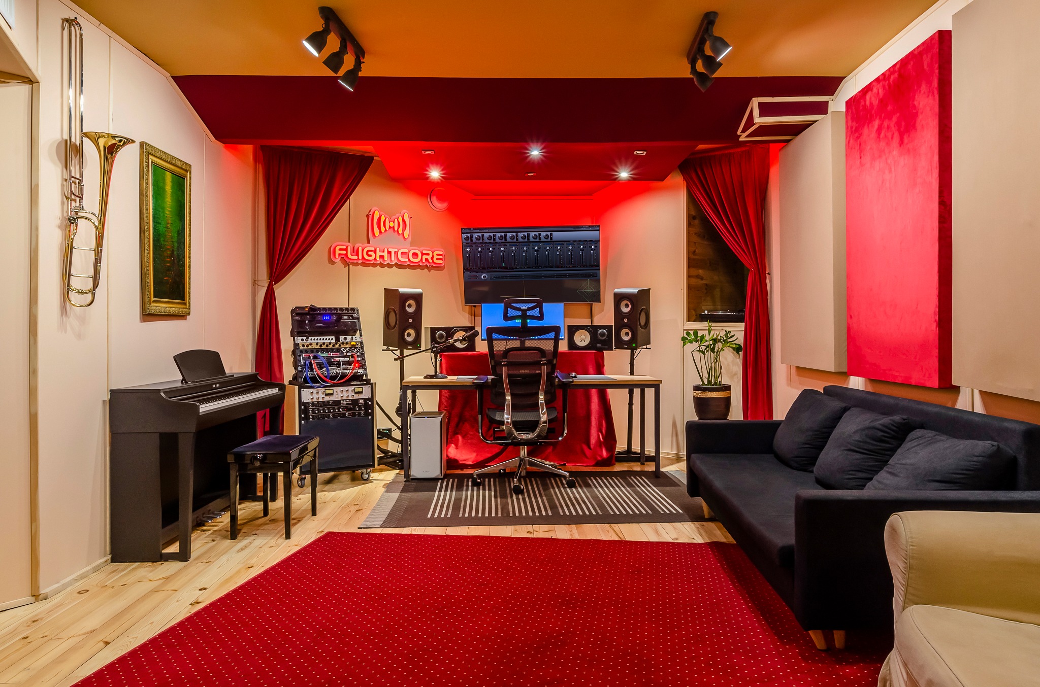 Profesjonalnie wyposażone pomieszczenie z pianinem, biurkiem z monitorem i głośnikami, sofą oraz neonem Flightcore, oświetlone czerwonym światłem.