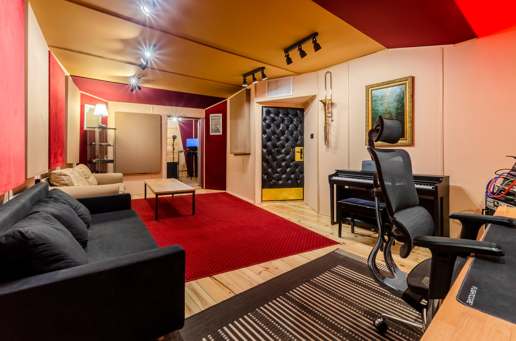 Wnętrze pokoju z pianinem, sofą, biurkiem i mikrofonem, prawdopodobnie pomieszczenie do nagrywania z akustycznymi panelami ściennymi i ozdobnym obrazem w złotej ramie.