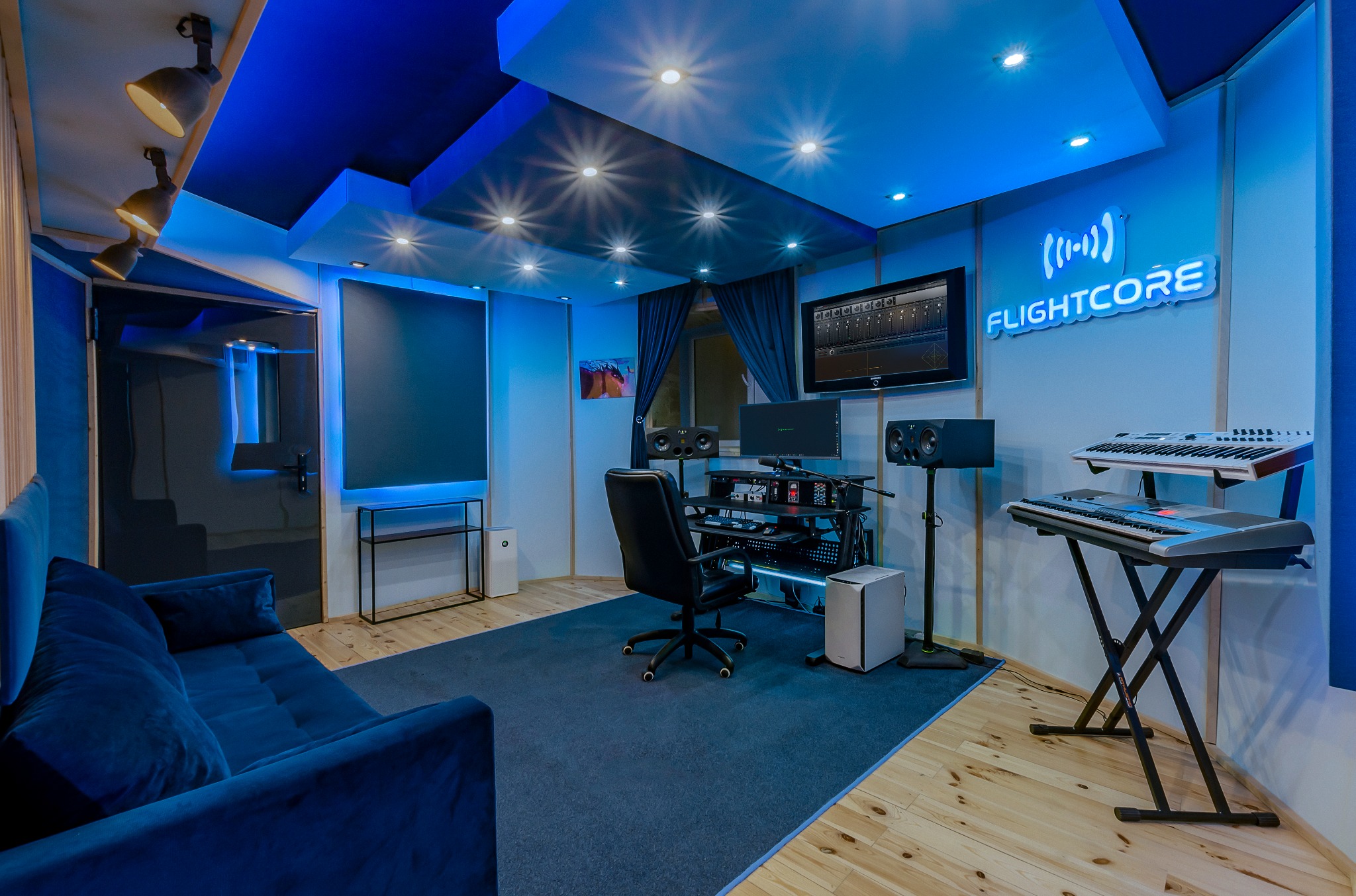 Nowoczesne pomieszczenie z niebieskim oświetleniem, sofą, biurkiem z monitorem, głośnikami, klawiaturą i neonem 'FLIGHTCORE' na ścianie, drewniana podłoga.