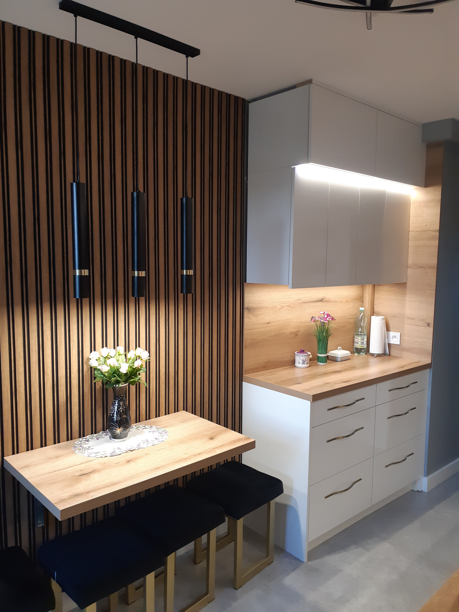 Nowoczesna kuchnia z drewnianymi lamelami na ścianie, podwieszanymi czarnymi lampami, blatem z jasnego drewna i białymi szafkami z podświetleniem LED, uzupełniona o stół z kwiatami i czarne pufy...
