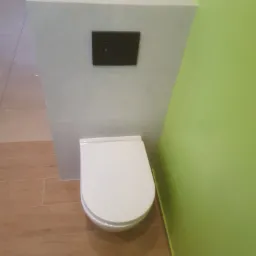 Zamknięta, biała toaleta wisząca ze spłuczką w kolorze czarnym, obudowana płytkami imitującymi marmur, na podłodze płytki imitujące drewno, ściana pomalowana na jasnozielono.