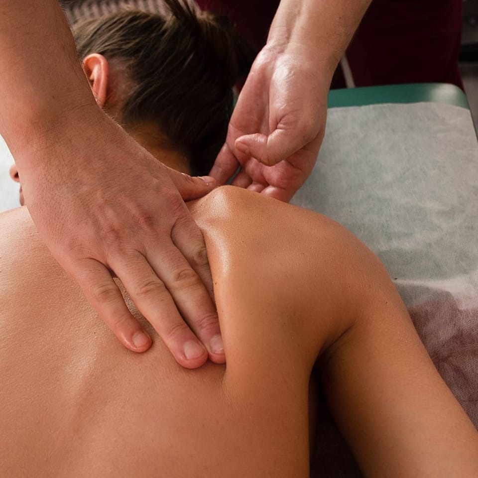 Terapeuta uciska dłonią mięśnie szyi i ramion pacjenta leżącego na stole pokrytym prześcieradłem.