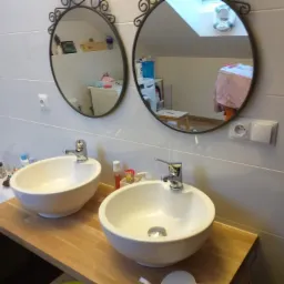 Dwie okrągłe umywalki nablatowe na drewnianym blacie w łazience z białymi płytkami, nad nimi dwa ozdobne lustra, w których odbija się okno dachowe i pralka.