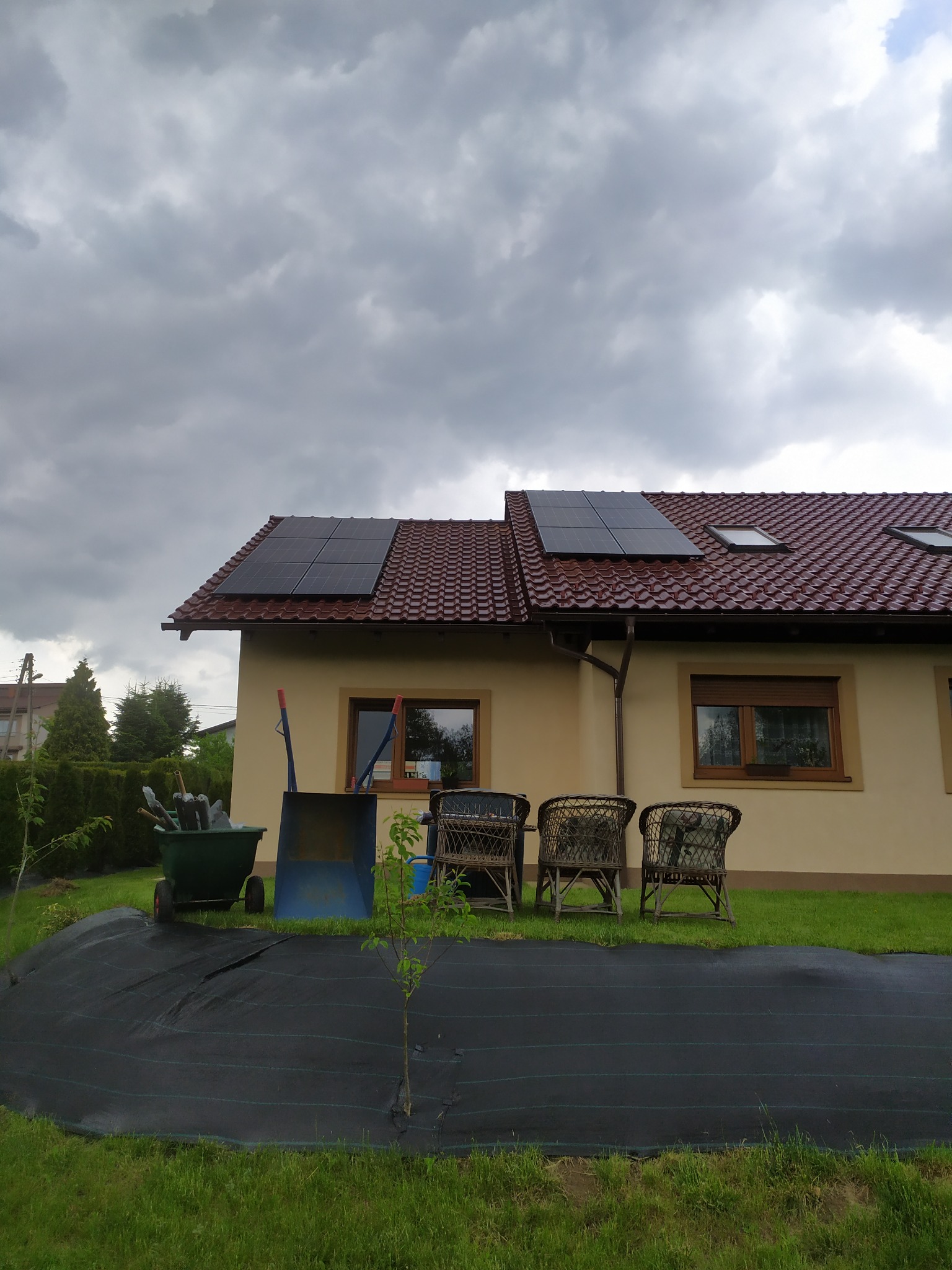 Dom jednorodzinny z czerwoną dachówką i panelami fotowoltaicznymi na dachu, widoczne narzędzia ogrodnicze i trzy wiklinowe krzesła na trawniku pod pochmurnym niebem.