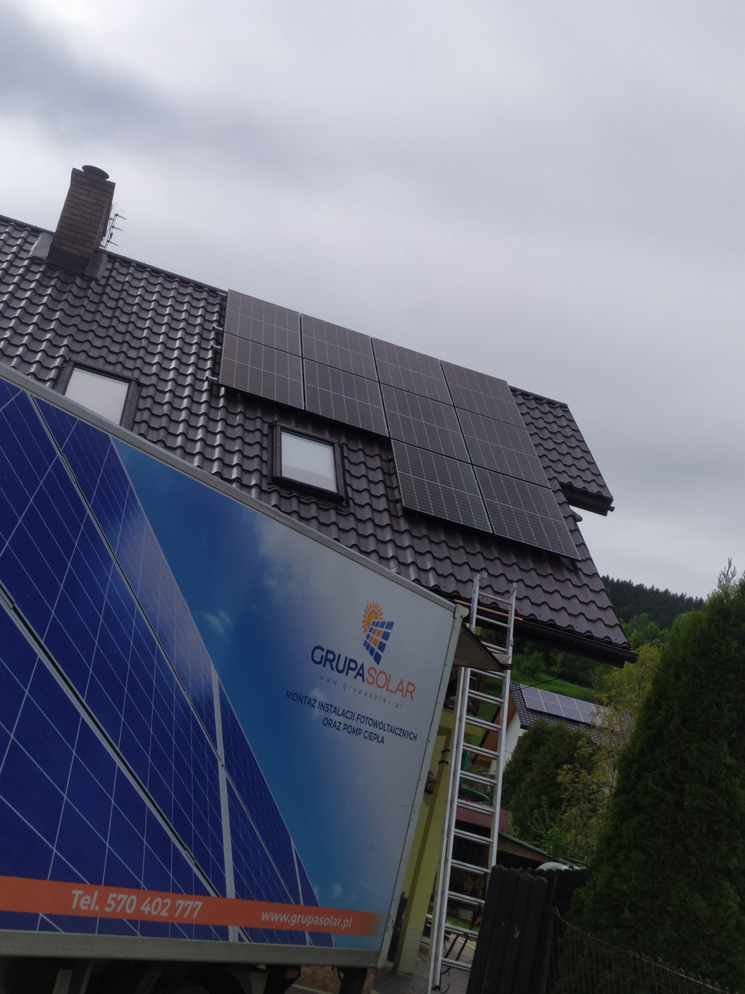 Montaż paneli słonecznych na brązowym dachu domu z widoczną drabiną i samochodem z logo firmy Grupa Solar