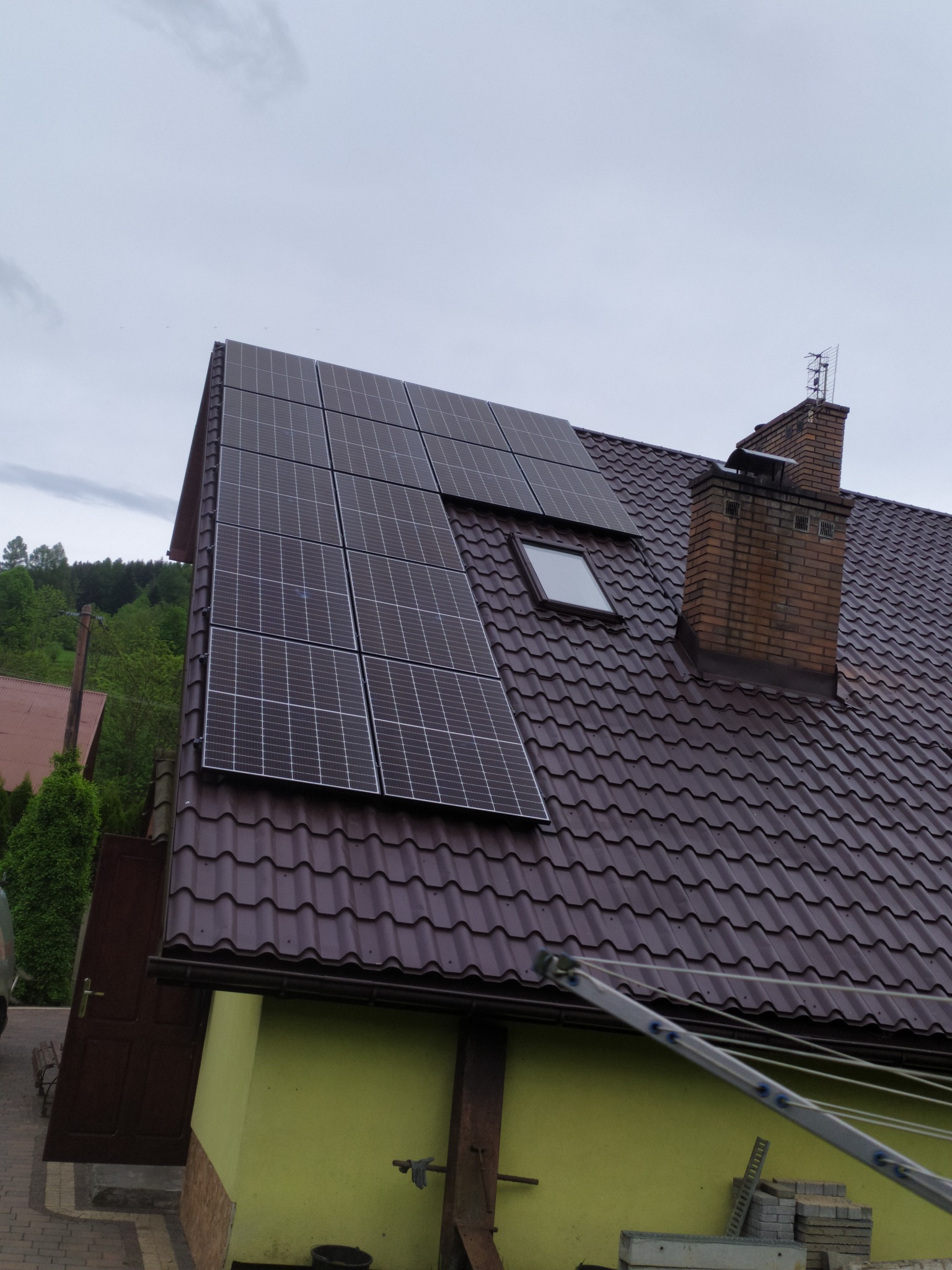 Panele słoneczne na brązowym dachu domu z ceglanym kominem i oknem dachowym; drabina oparta o ścianę budynku w tle.