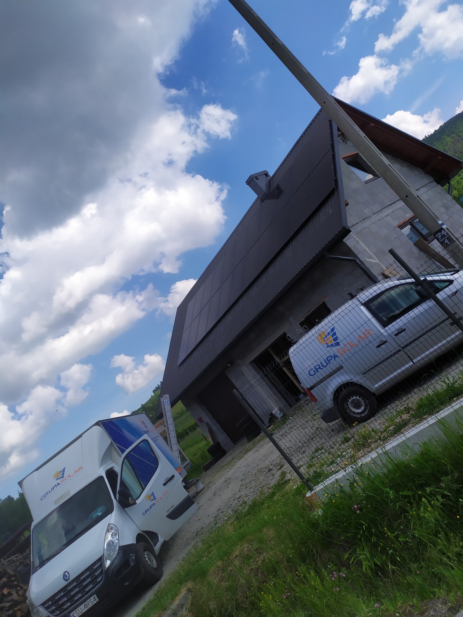 Montaż paneli fotowoltaicznych na dachu domu jednorodzinnego, widoczne dwa samochody dostawcze firmy Grupa Solar na tle błękitnego nieba z chmurami.