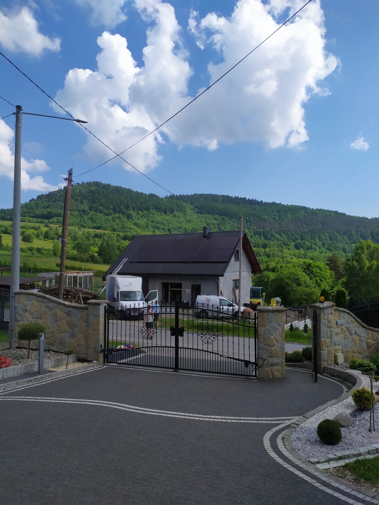 Montaż paneli na dachu domu jednorodzinnego z widocznymi dwoma samochodami dostawczymi i pracownikami na drabinie, w tle zalesione wzgórze.