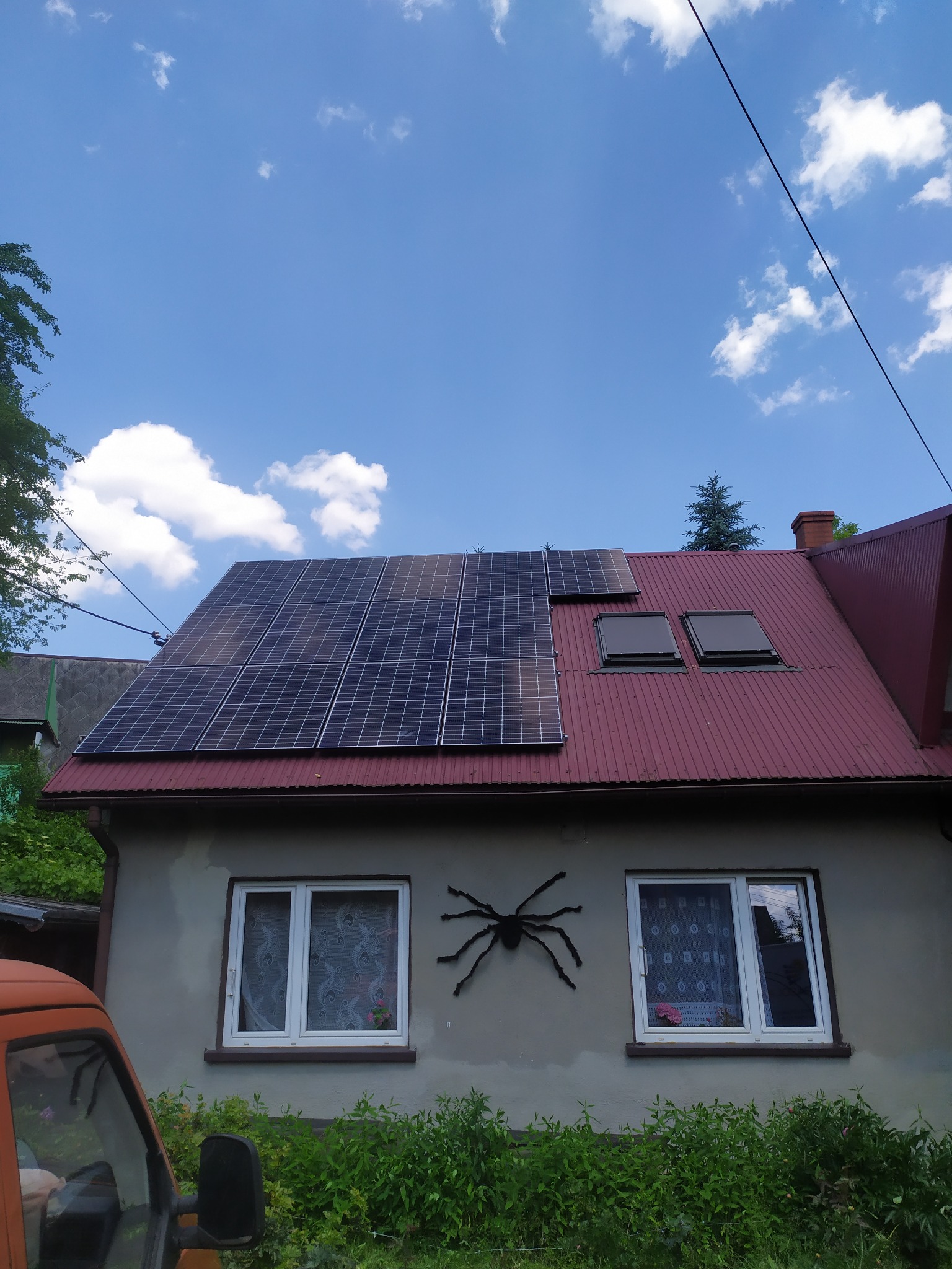 Dom jednorodzinny z panelami fotowoltaicznymi na czerwonym dachu, ozdobiony dekoracją w kształcie pająka na elewacji.