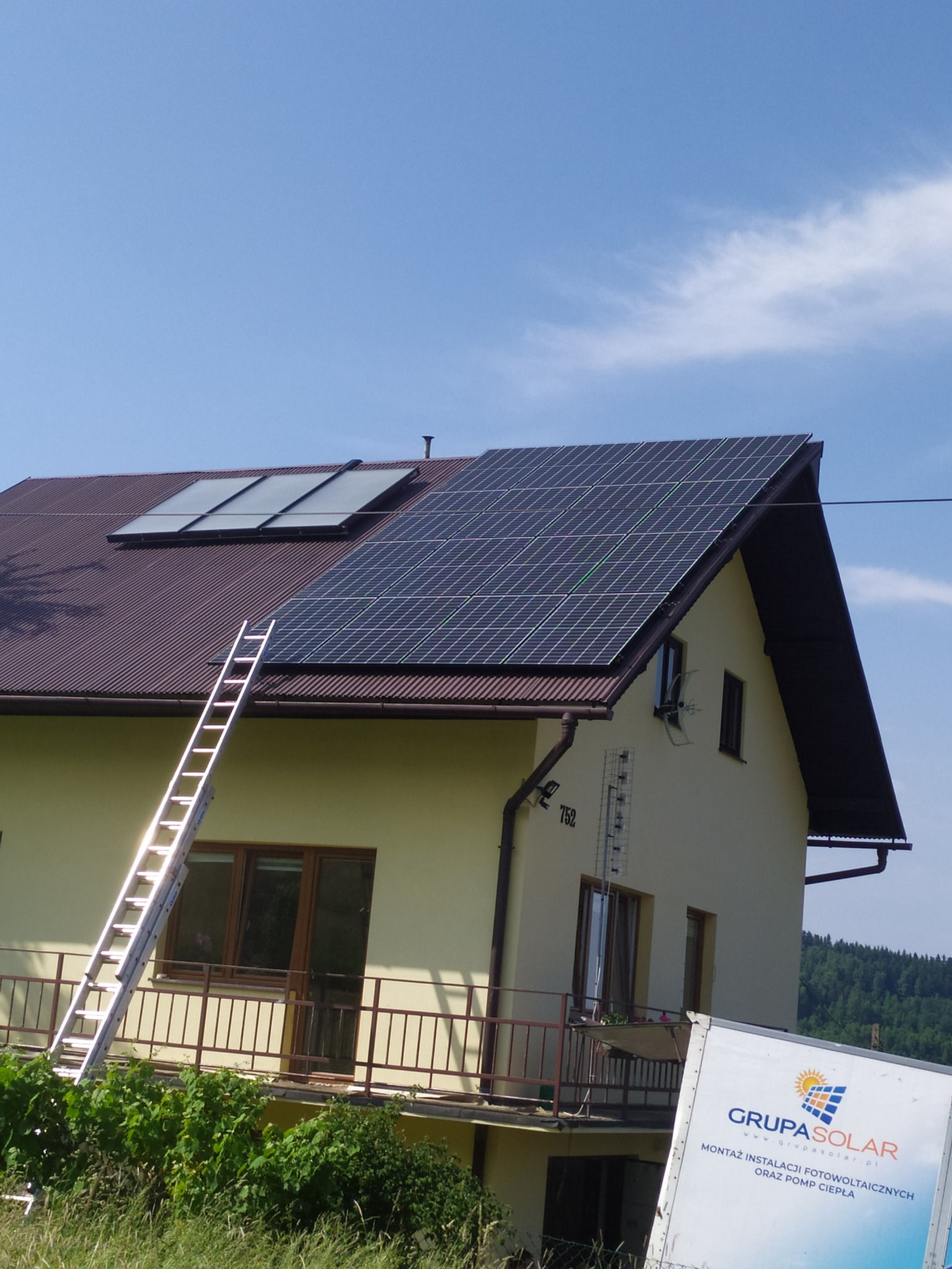 Dom z brązowym dachem, na którym zamontowane są panele słoneczne i kolektory słoneczne, z przystawioną drabiną i widocznym samochodem dostawczym z logo firmy instalującej.