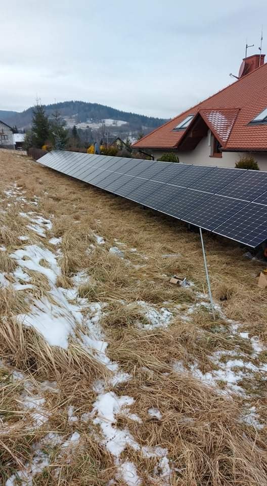 Długi rząd paneli słonecznych zamontowany na stelażach na zboczu porośniętym suchą trawą i resztkami śniegu, dom z czerwoną dachówką w tle.