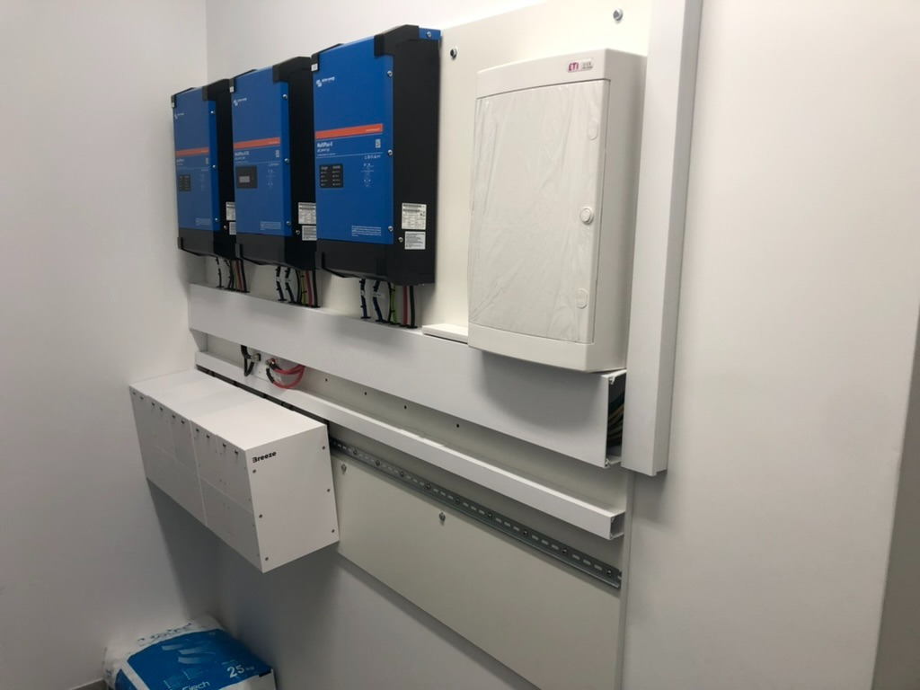 Instalacja z trzema niebieskimi falownikami Victron Energy MultiPlus-II, białą skrzynką elektryczną i skrzynką Breeze na białej ścianie, widoczne okablowanie.