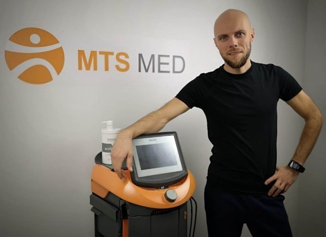 Mężczyzna z brodą opiera się o pomarańczowe urządzenie medyczne z ekranem dotykowym, na tle logo MTS MED.