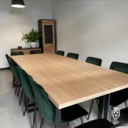 Jasna sala konferencyjna z dużym drewnianym stołem i krzesłami z ciemnozielonego weluru, z widoczną szafką z rośliną w tle.