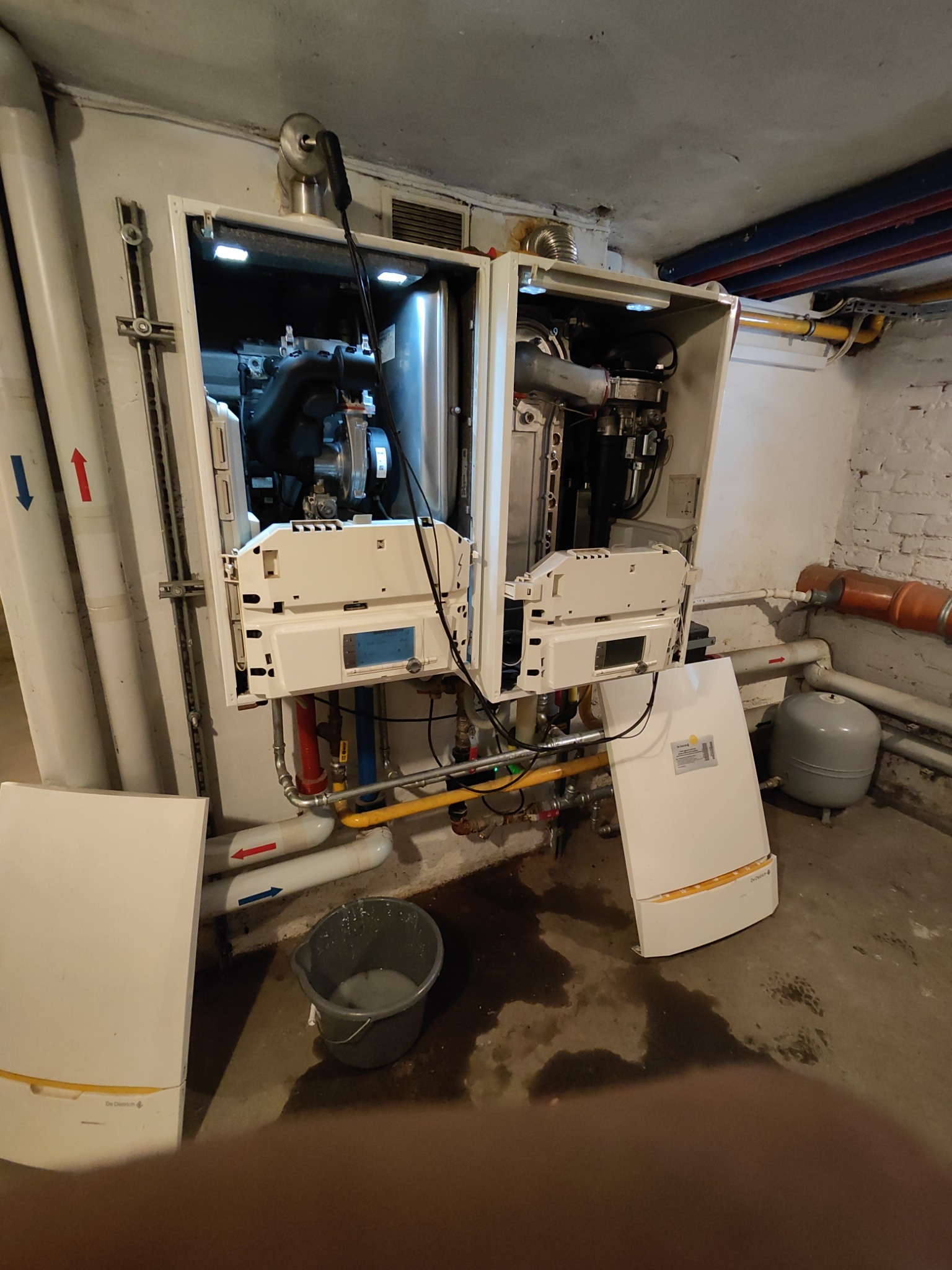 Otwarty piec gazowy De Dietrich w trakcie montażu lub serwisu, widoczne rury, instalacja, wiadro z wodą i mokra podłoga w piwnicy.