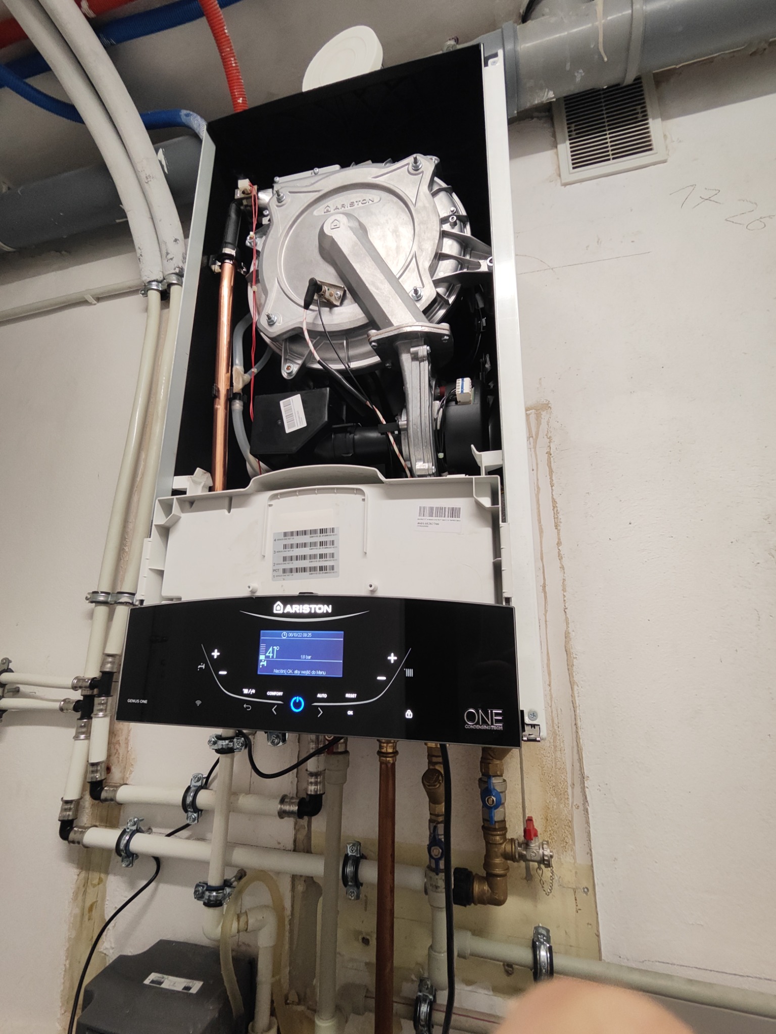 Otwarty piec gazowy Ariston Genus One z widocznymi elementami wewnętrznymi i podłączonymi rurami miedzianymi oraz plastikowymi, zamontowany na ścianie z widocznymi śladami po instalacji.