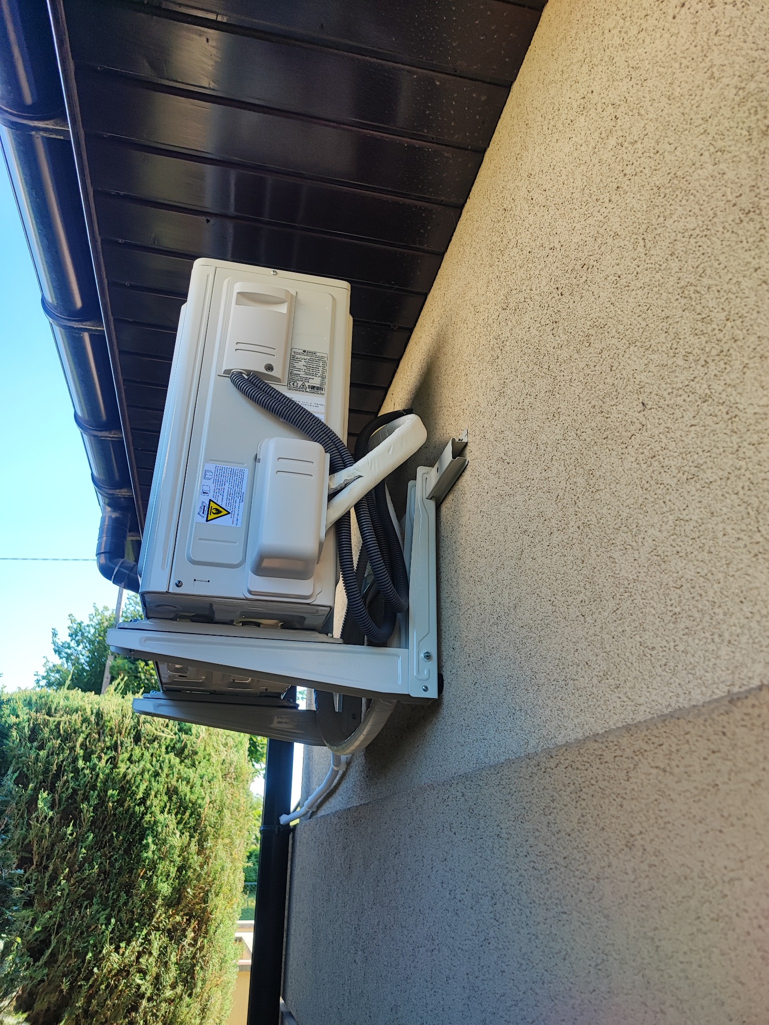 Zewnętrzna jednostka klimatyzacji zamontowana na ścianie budynku, widoczne rury odprowadzające skropliny i przewody elektryczne.