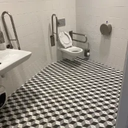 Toaleta dla niepełnosprawnych z białymi płytkami ściennymi, poręczami ze stali nierdzewnej i geometryczną, czarno-biało-szarą podłogą z efektem 3D.