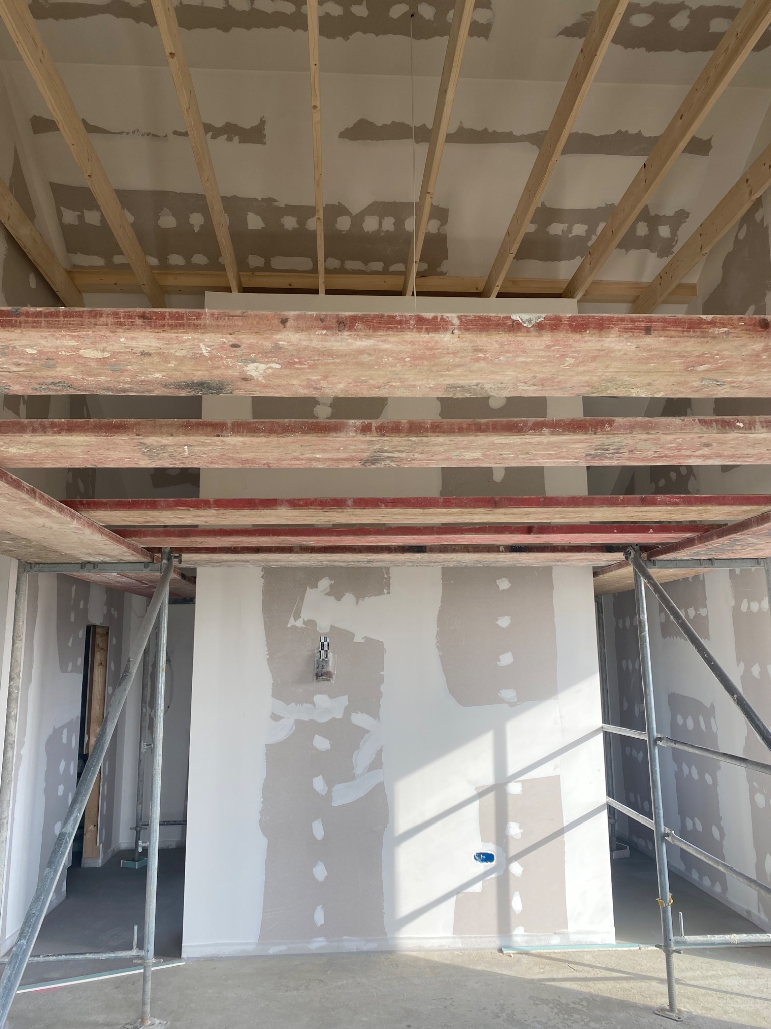 Wnętrze w trakcie szpachlowania ścian i sufitu poddasza, widoczne drewniane krokwie stropu, metalowe rusztowanie i niedokończone gładzie gipsowe.