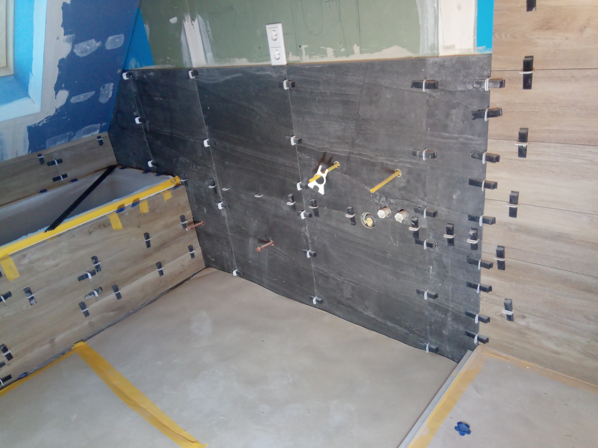 Ściana łazienki w trakcie układania płytek: ciemnoszare płytki imitujące kamień sąsiadują z jasnymi, drewnopodobnymi, widoczne krzyżyki dystansowe, przygotowane otwory na instalację wodną...