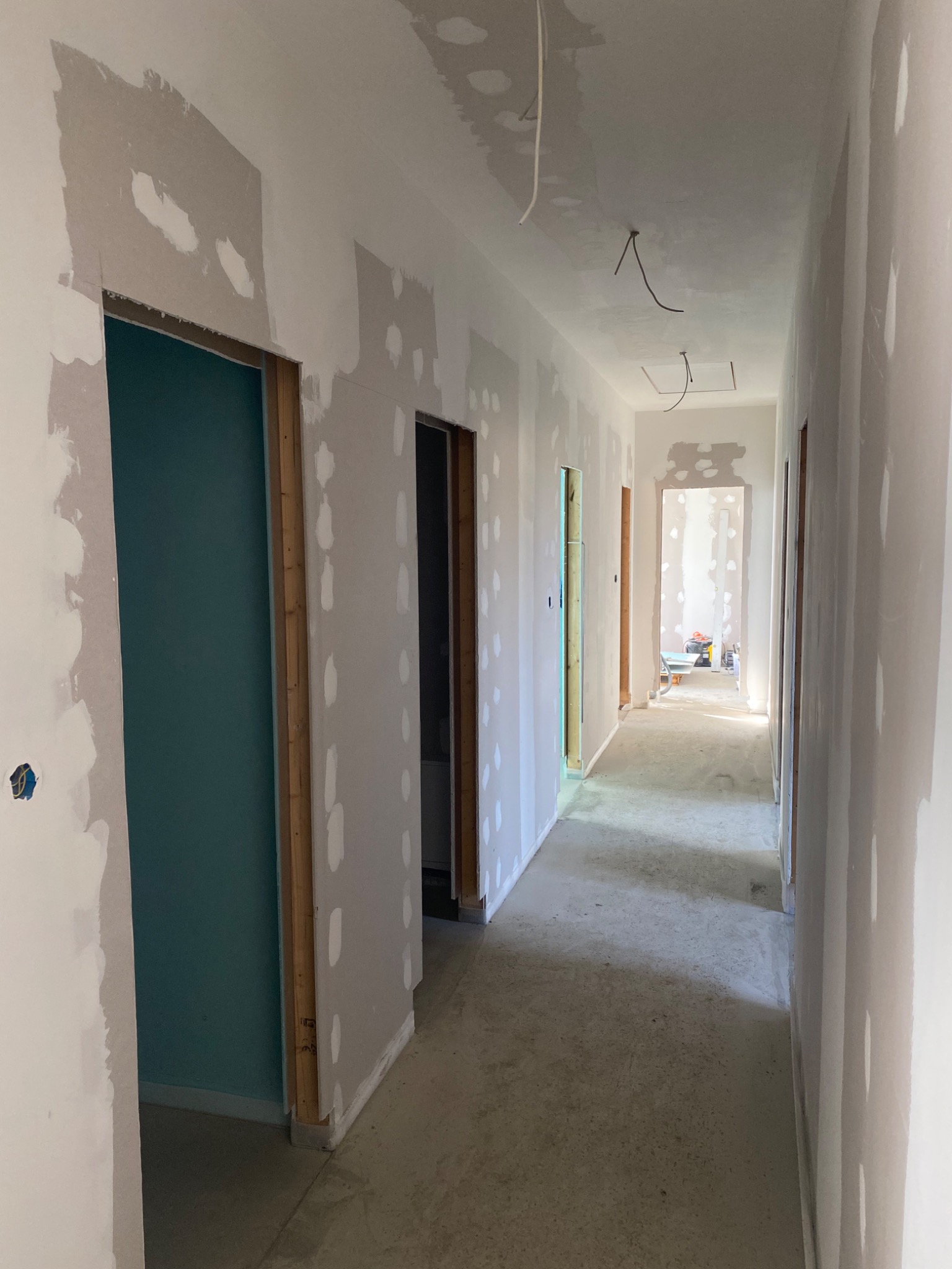 Korytarz w trakcie prac wykończeniowych: ściany częściowo pokryte gładzią, widoczne kable wystające z sufitu, surowa podłoga betonowa, otwory drzwiowe bez ościeżnic, w oddali wózek budowlany.